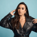 Demi Lovato mengenakan jaket kulit hitam berpose di depan latar biru.